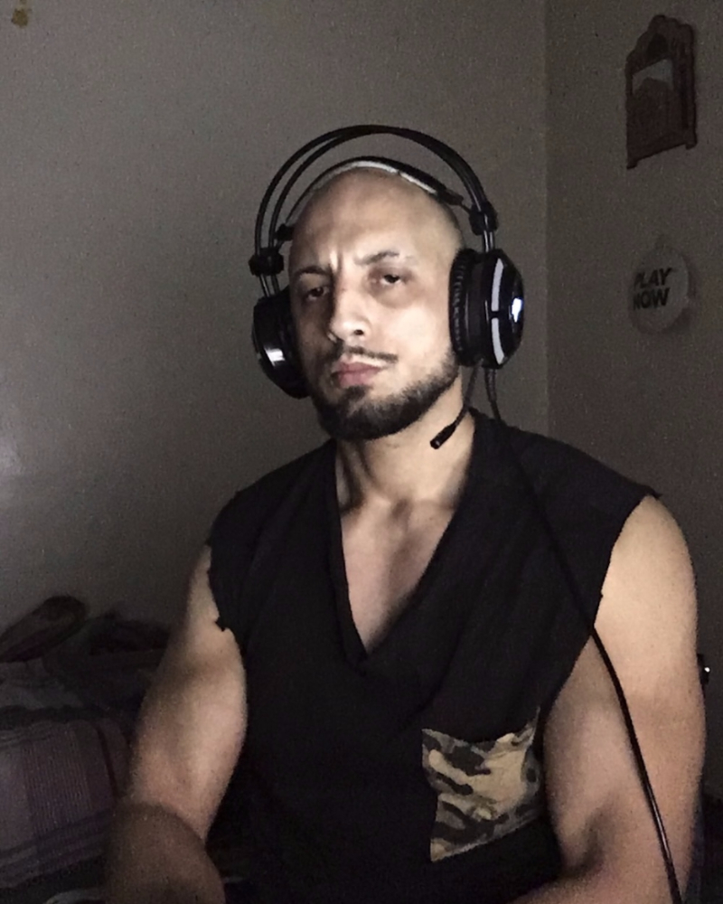 Portrait de Hamza Anis Chellouche, portant un casque audio avec micro, assis dans une pièce sombre. Il porte un t-shirt noir sans manches avec une poche à motif camouflage.