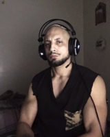 Portrait de Hamza Anis Chellouche, portant un casque audio avec micro, assis dans une pièce sombre. Il porte un t-shirt noir sans manches avec une poche à motif camouflage.