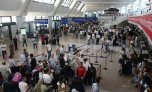 Hall des départs bondé de l'aéroport Houari-Boumediene d'Alger en été, voyageurs en attente aux comptoirs d'enregistrement.