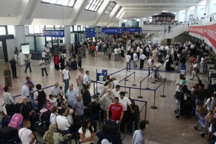 Hall des départs bondé de l'aéroport Houari-Boumediene d'Alger en été, voyageurs en attente aux comptoirs d'enregistrement.