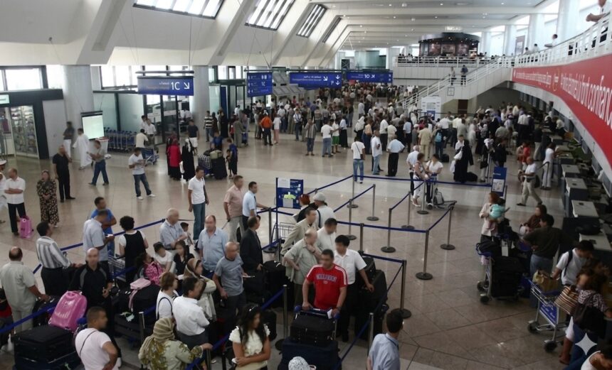 Hall des départs bondé de l'aéroport Houari-Boumediene d'Alger en été, voyageurs en attente aux comptoirs d'enregistrement.
