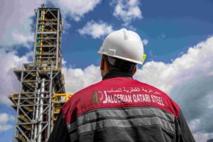 Employé de dos portant une veste de travail floquée du logo Algerian Qatari Steel, devant une tour du complexe sidérurgique de Bellara