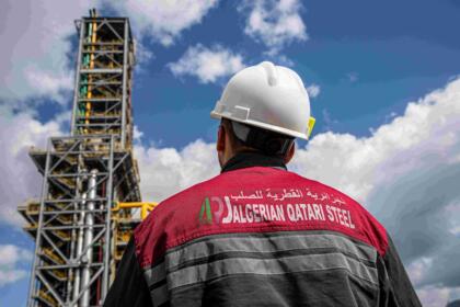 Employé de dos portant une veste de travail floquée du logo Algerian Qatari Steel, devant une tour du complexe sidérurgique de Bellara