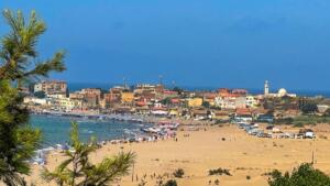 Vue depuis les dunes de Benyounes sur la plage de Zemmouri El Bahri, animée de baigneurs et de parasols, avec le village et le minaret de sa mosquée en arrière-plan, face à la Méditerranée