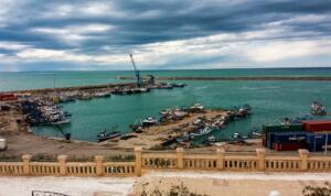 Vue sur le port de pêche de Mostaganem avec des bateaux de pêche, des quais et des conteneurs, sous un ciel nuageux.