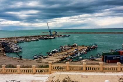 Vue sur le port de pêche de Mostaganem avec des bateaux de pêche, des quais et des conteneurs, sous un ciel nuageux.