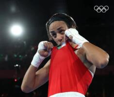 Imane Khelif, gants de boxe levés, portant à ses lèvres le maillot floqué du drapeau algérien, anneaux olympiques en arrière-plan.