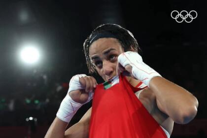 Imane Khelif, gants de boxe levés, portant à ses lèvres le maillot floqué du drapeau algérien, anneaux olympiques en arrière-plan.