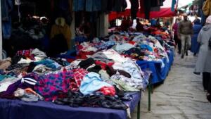 Marché de vêtements de seconde main en plein air à Alger, avec des piles de vêtements colorés sur des tables et des passants dans une ruelle pavée.
