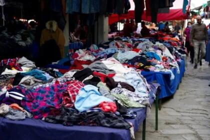 Marché de vêtements de seconde main en plein air à Alger, avec des piles de vêtements colorés sur des tables et des passants dans une ruelle pavée.