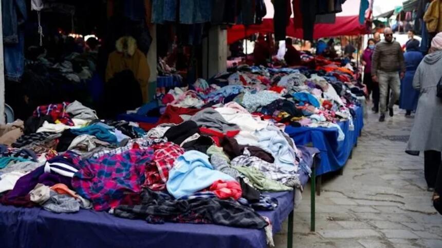 Marché de vêtements de seconde main en plein air à Alger, avec des piles de vêtements colorés sur des tables et des passants dans une ruelle pavée.