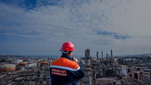 Un technicien Sonatrach en tenue orange et bleue, casque rouge, observe un complexe pétrolier et gazier en bord de mer.