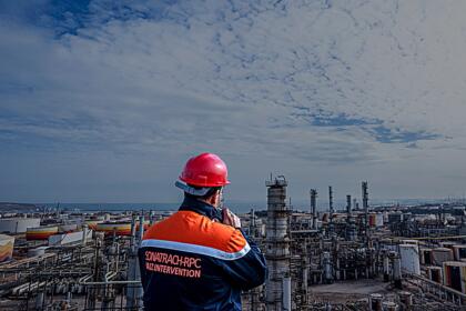 Un technicien Sonatrach en tenue orange et bleue, casque rouge, observe un complexe pétrolier et gazier en bord de mer.