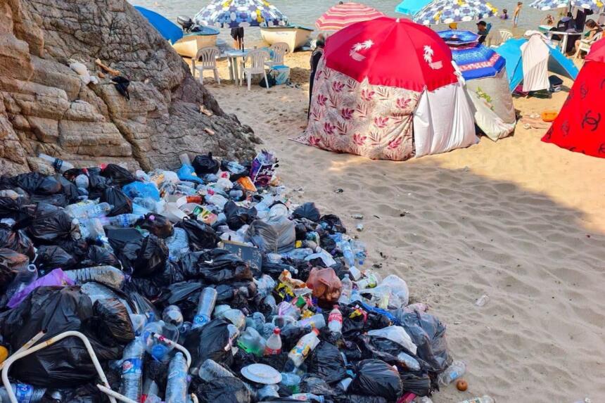 Tas de sacs-poubelle et bouteilles en plastique sur une plage, avec parasols et baigneurs en arrière-plan.