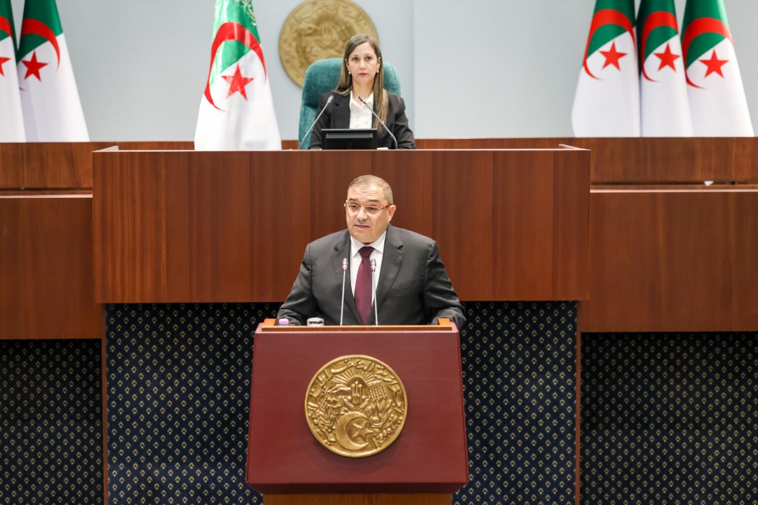 Le ministre de la Justice, Lotfi Boudjemaa, s'exprime à la tribune de l'Assemblée populaire nationale, entre des drapeaux algériens.