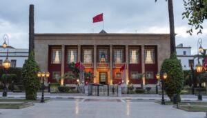 Façade du Parlement marocain à Rabat, avec ses colonnes, ses drapeaux du Maroc et ses lampadaires allumés, sous un ciel nuageux