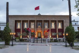 Façade du Parlement marocain à Rabat, avec ses colonnes, ses drapeaux du Maroc et ses lampadaires allumés, sous un ciel nuageux