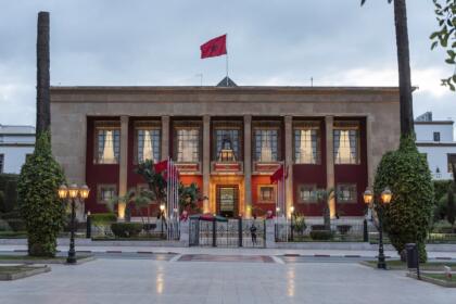 Façade du Parlement marocain à Rabat, avec ses colonnes, ses drapeaux du Maroc et ses lampadaires allumés, sous un ciel nuageux