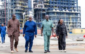 Quatre personnes marchent côte à côte sur une aire industrielle, devant les structures métalliques d'une raffinerie de pétrole. Un conteneur jaune est visible à droite.