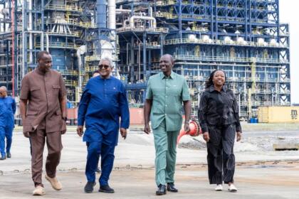 Quatre personnes marchent côte à côte sur une aire industrielle, devant les structures métalliques d'une raffinerie de pétrole. Un conteneur jaune est visible à droite.