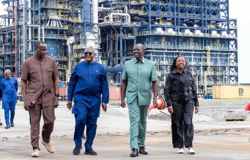 Quatre personnes marchent côte à côte sur une aire industrielle, devant les structures métalliques d'une raffinerie de pétrole. Un conteneur jaune est visible à droite.