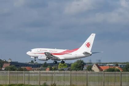 Avion de ligne blanc et rouge aux couleurs d'Air Algérie en phase d'atterrissage, train sorti, au-dessus d'une clôture d'aéroport bordée de maisons.