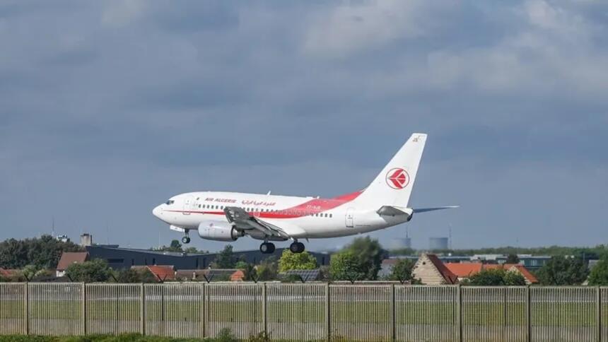 Avion de ligne blanc et rouge aux couleurs d'Air Algérie en phase d'atterrissage, train sorti, au-dessus d'une clôture d'aéroport bordée de maisons.