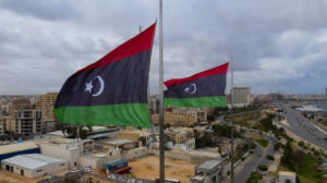 Deux drapeaux libyens (bandes rouge, noire et verte, croissant et étoile blancs) flottent au vent en haut de mâts métalliques. En contrebas, une ville côtière d'immeubles beiges et de hangars est longée par une avenue et bordée par la mer. Le ciel est gris et nuageux.