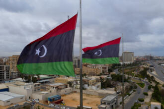Deux drapeaux libyens (bandes rouge, noire et verte, croissant et étoile blancs) flottent au vent en haut de mâts métalliques. En contrebas, une ville côtière d'immeubles beiges et de hangars est longée par une avenue et bordée par la mer. Le ciel est gris et nuageux.