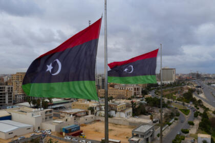 Deux drapeaux libyens (bandes rouge, noire et verte, croissant et étoile blancs) flottent au vent en haut de mâts métalliques. En contrebas, une ville côtière d'immeubles beiges et de hangars est longée par une avenue et bordée par la mer. Le ciel est gris et nuageux.
