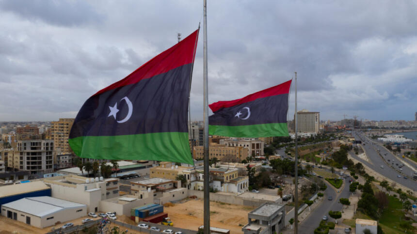 Deux drapeaux libyens (bandes rouge, noire et verte, croissant et étoile blancs) flottent au vent en haut de mâts métalliques. En contrebas, une ville côtière d'immeubles beiges et de hangars est longée par une avenue et bordée par la mer. Le ciel est gris et nuageux.