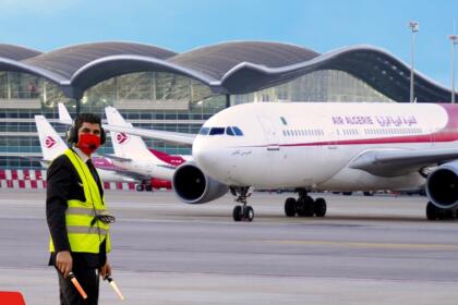 Avion Airbus A330 d'Air Algérie sur le tarmac de l'aéroport Houari Boumediene d'Alger, agent au sol en gilet jaune au premier plan, terminal à toiture ondulée en arrière-plan.