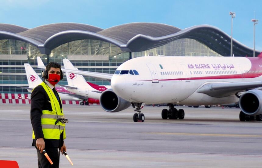 Avion Airbus A330 d'Air Algérie sur le tarmac de l'aéroport Houari Boumediene d'Alger, agent au sol en gilet jaune au premier plan, terminal à toiture ondulée en arrière-plan.