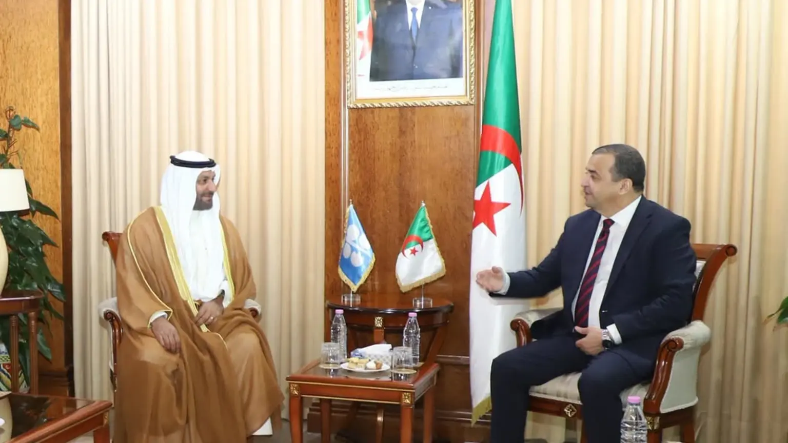 Mohamed Arkab, assis à droite, s'entretient avec Haitham Al-Ghais, en tenue traditionnelle du Golfe, dans un salon lambrissé du ministère des Hydrocarbures ; quatre responsables algériens en costume sont assis à gauche, sous le portrait du président et le drapeau algérien.