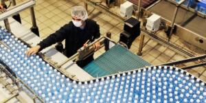 Vue plongeante sur une ligne de production industrielle où un ouvrier portant un masque et une charlotte vérifie un tapis roulant rempli de bouteilles d'eau en plastique bleu.