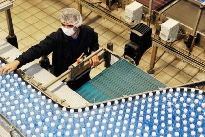 Vue plongeante sur une ligne de production industrielle où un ouvrier portant un masque et une charlotte vérifie un tapis roulant rempli de bouteilles d'eau en plastique bleu.