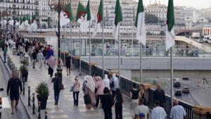 Des passants marchent sur une large promenade piétonne d'Alger, bordée de mâts portant des drapeaux algériens, avec des immeubles blancs de style colonial en arrière-plan.
