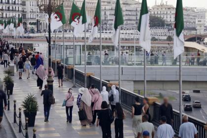 Des passants marchent sur une large promenade piétonne d'Alger, bordée de mâts portant des drapeaux algériens, avec des immeubles blancs de style colonial en arrière-plan.