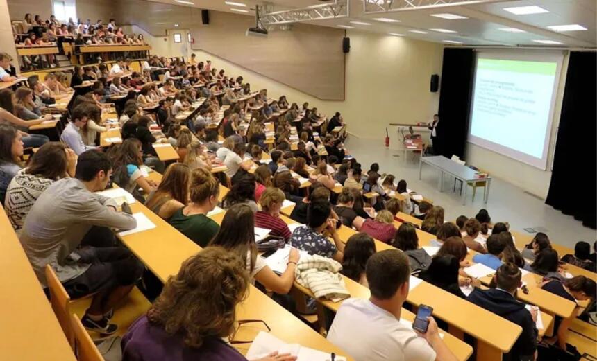 Amphithéâtre universitaire bondé, avec des étudiants assis sur des rangées de bancs en bois pendant un cours.