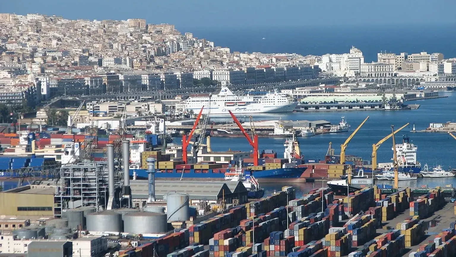 Vue du port d'Alger avec des conteneurs empilés, des grues de déchargement, un cargo à quai et la ville en arrière-plan