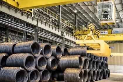 Bobines de fil machine en acier empilées dans une usine sidérurgique, avec un pont roulant industriel en arrière-plan.