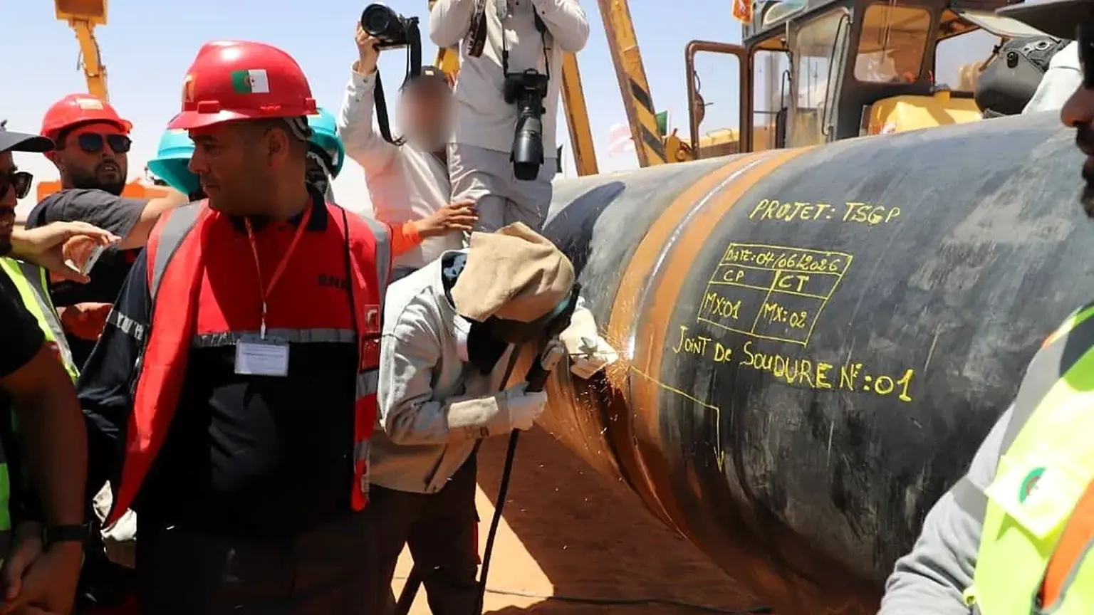 Un soudeur casqué réalise une soudure sur un gros tube noir du gazoduc transsaharien. Le tube porte l'inscription à la craie « Projet TSGP, 04/06/2026, joint de soudure n° 01 ». Il est entouré d'ouvriers en casques rouges portant le drapeau algérien et de photographes, sur un chantier sablonneux, avec des engins en arrière-plan.