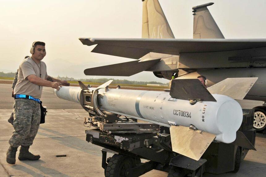 Technicien militaire américain en tenue de treillis installant un missile AIM-120 sous l'aile d'un avion de chasse sur un tarmac.