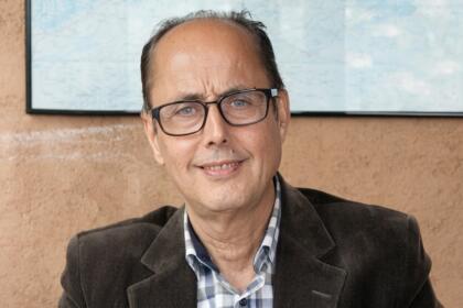 Portrait de l'économiste Omar Berkouk, souriant, lunettes et veste en velours côtelé, devant une carte murale.