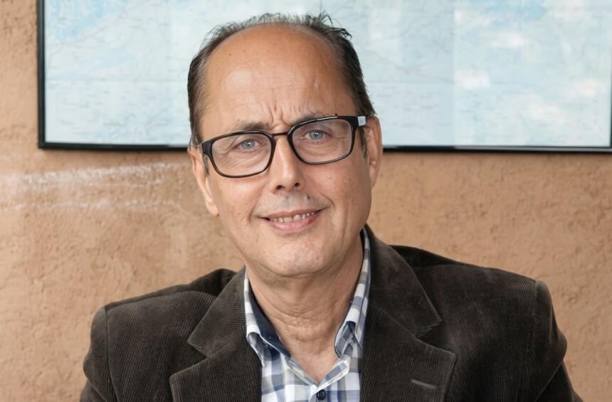 Portrait de l'économiste Omar Berkouk, souriant, lunettes et veste en velours côtelé, devant une carte murale.