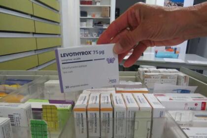 Main de pharmacienne saisissant une boîte de Levothyrox 75 µg dans un rayonnage de pharmacie, entourée d'autres boîtes de Levothyroxine.