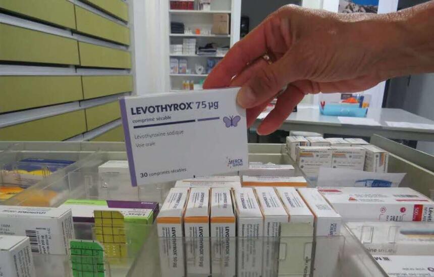 Main de pharmacienne saisissant une boîte de Levothyrox 75 µg dans un rayonnage de pharmacie, entourée d'autres boîtes de Levothyroxine.