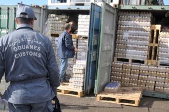 عون من الجمارك الجزائرية يرتدي زيًا رسميًا مكتوبًا عليه "الجمارك الجزائرية – Douanes – Customs"، يقف أمام حاويتين مفتوحتين محمّلتين بمنصّات من العبوات البلاستيكية المغلّفة، بينما يتفقد رجل آخر الشحنة داخل إحدى الحاويات.
