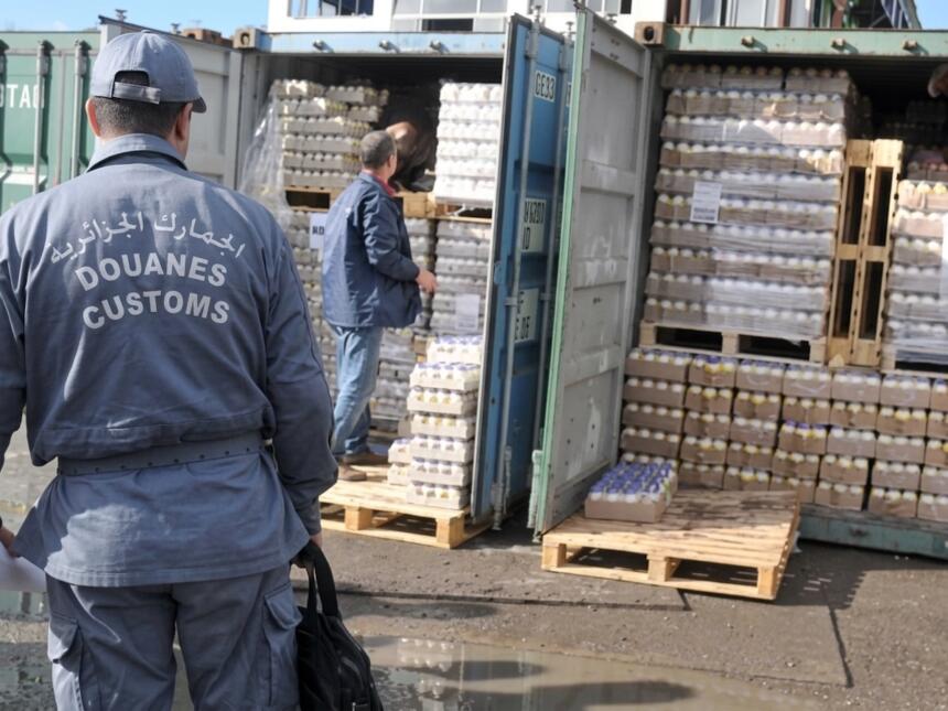 عون من الجمارك الجزائرية يرتدي زيًا رسميًا مكتوبًا عليه "الجمارك الجزائرية – Douanes – Customs"، يقف أمام حاويتين مفتوحتين محمّلتين بمنصّات من العبوات البلاستيكية المغلّفة، بينما يتفقد رجل آخر الشحنة داخل إحدى الحاويات.