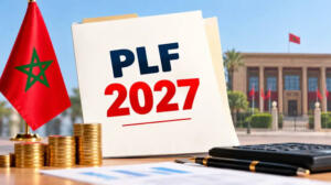 Un document portant la mention « PLF 2027 » posé à côté d’un drapeau marocain, de piles de pièces dorées, d’une calculatrice et d’un stylo, devant un bâtiment officiel.